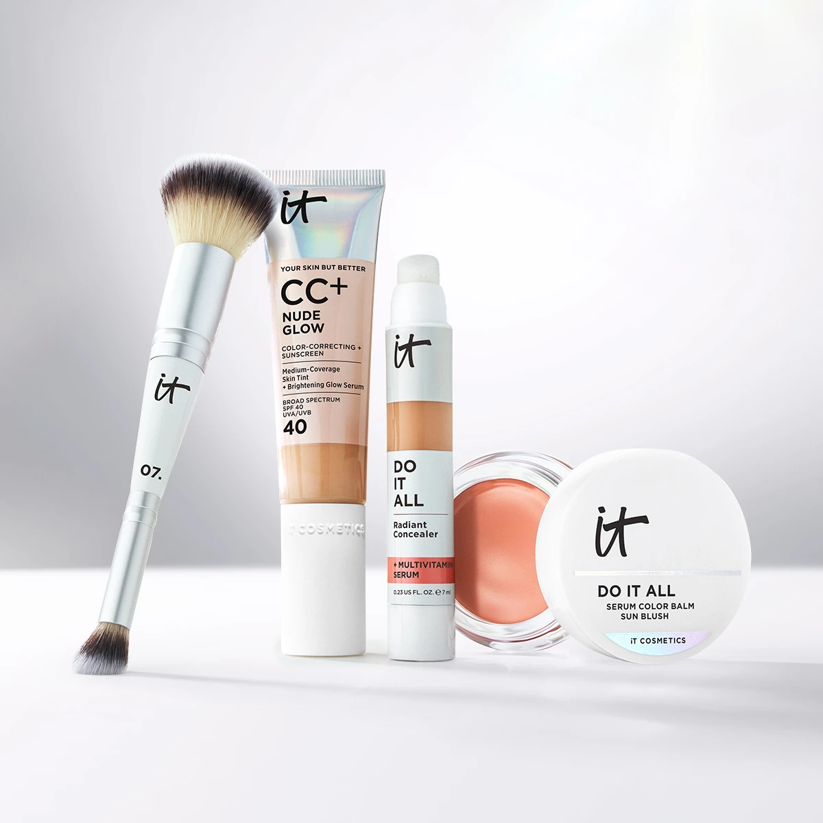 IT Cosmetics -meikkituotteita vaalealla taustalla, mukana meikkisivellin, CC+ Nude Glow, Do It All Radiant Concealer ja Do It All Serum Color Balm Sun Blush.
