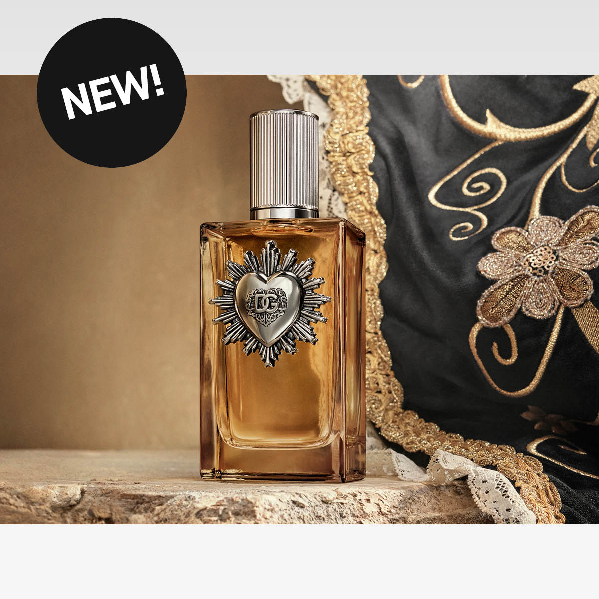 Parfymeflaske fra Dolce & Gabbana med metallhjerte på fronten, med svart stoff med gullbroderier i bakgrunnen. Sticker med teksten "NEW!" øverst til venstre.