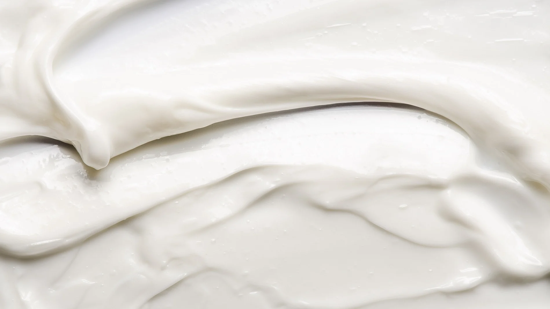 Närbild av en vit hudvårdskräm med mjuk, krämig konsistens utsmetad i böljande rörelser. Texturbild som framhäver en återfuktande ansiktskräm eller body lotion med glansig finish.