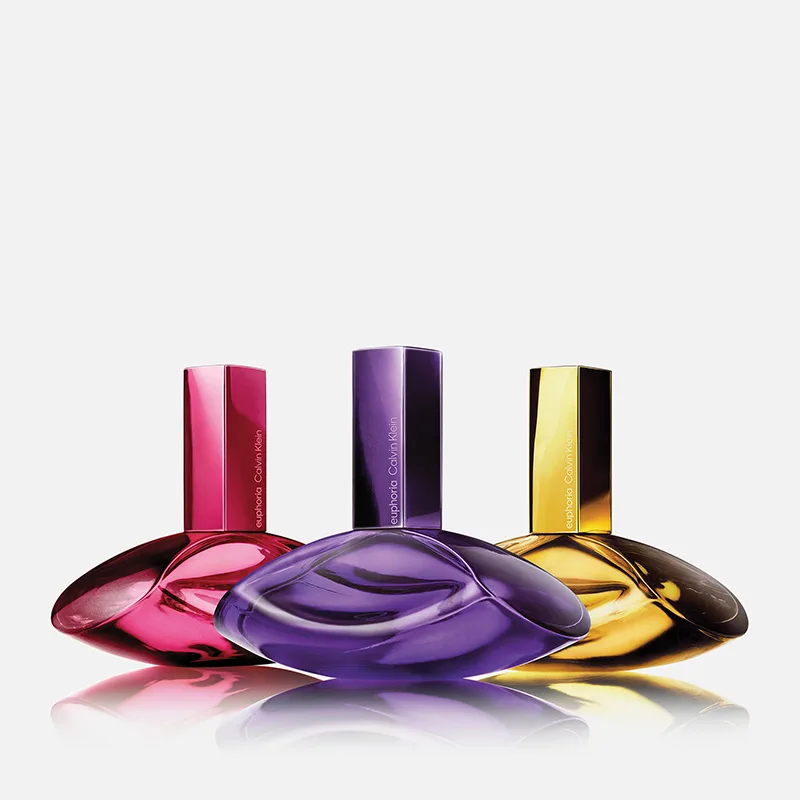 Tre fargede parfymeflasker fra Calvin Klein Euphoria står ved siden av hverandre på lys bakgrunn i rosa, lilla og gull med teksten "euphoria Calvin Klein" på korkene.