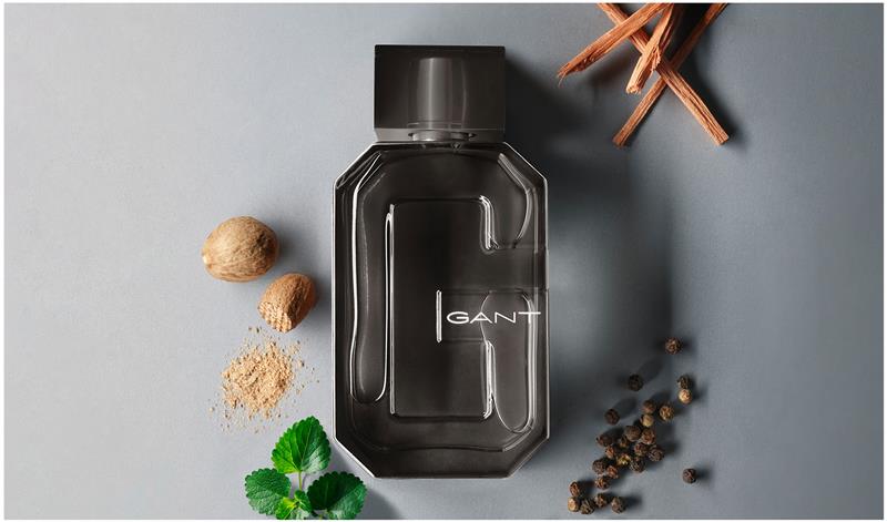 Mørk glassflaske med parfyme fra GANT plassert på en grå overflate, omgitt av krydder som pepperkorn, muskat og friske grønne blader. GANT-logoen er tydelig synlig på flasken med et moderne, kantet design.