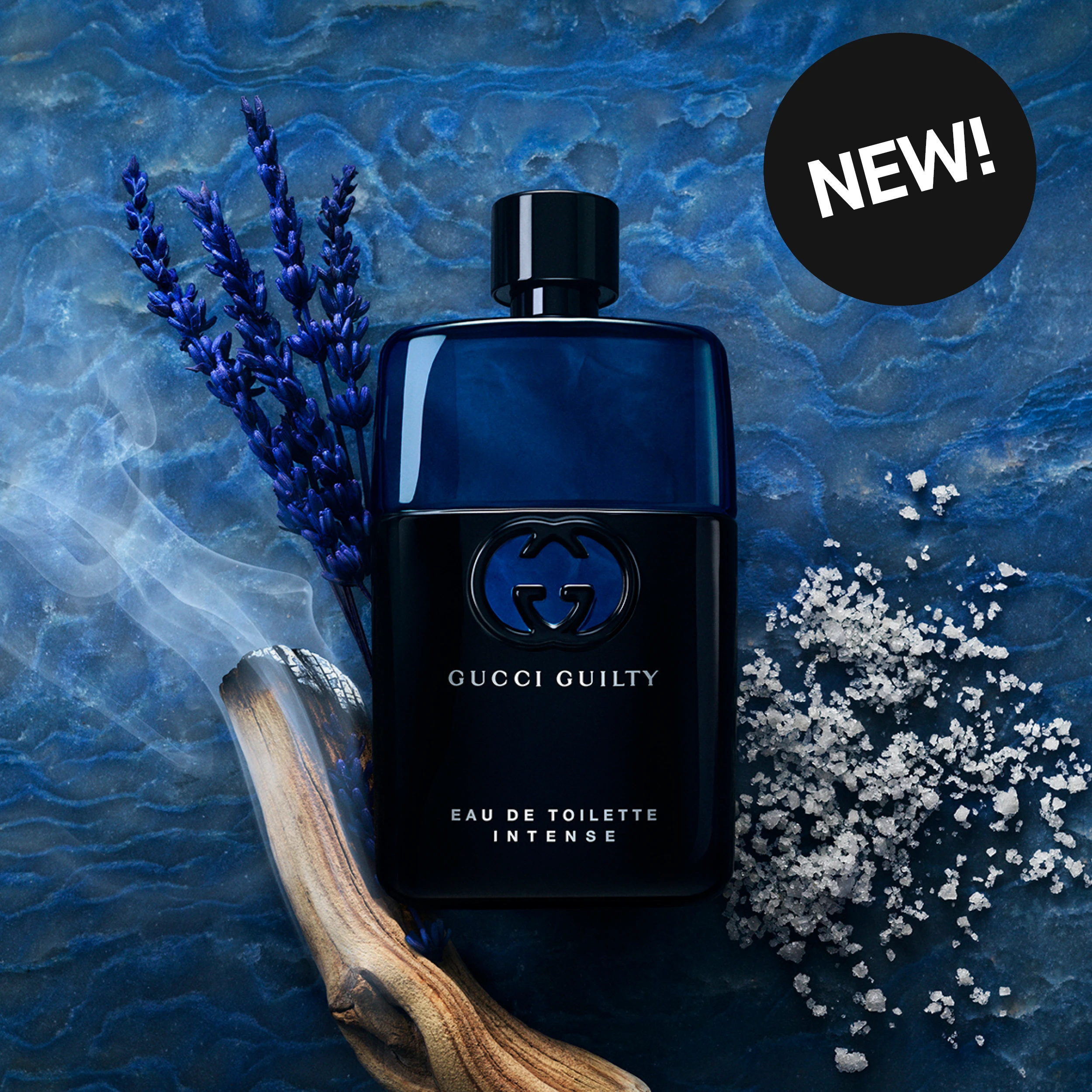 NEW! Gucci Guilty Eau de Toilette Intense-parfyme i en mørkeblå og svart flaske, omgitt av blå blomster, tre, røyk og hvite krystaller mot en blå bakgrunn.