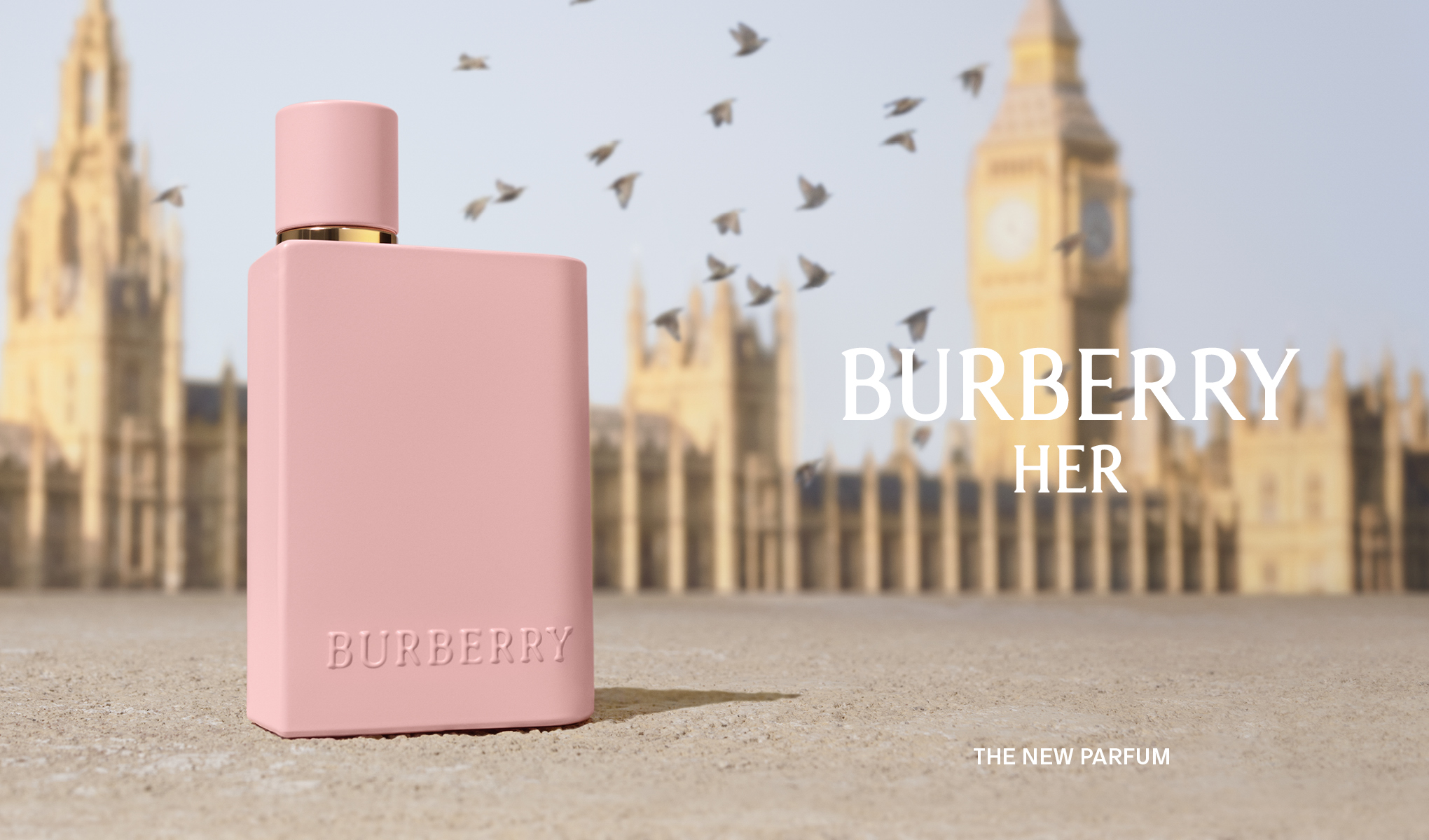 Burberry HER - den nye parfymen. En rosa Burberry HER-parfymeflaske står foran Londons ikoniske bygninger, inkludert Big Ben og Westminsterpalasset. Fugler flyr i bakgrunnen.