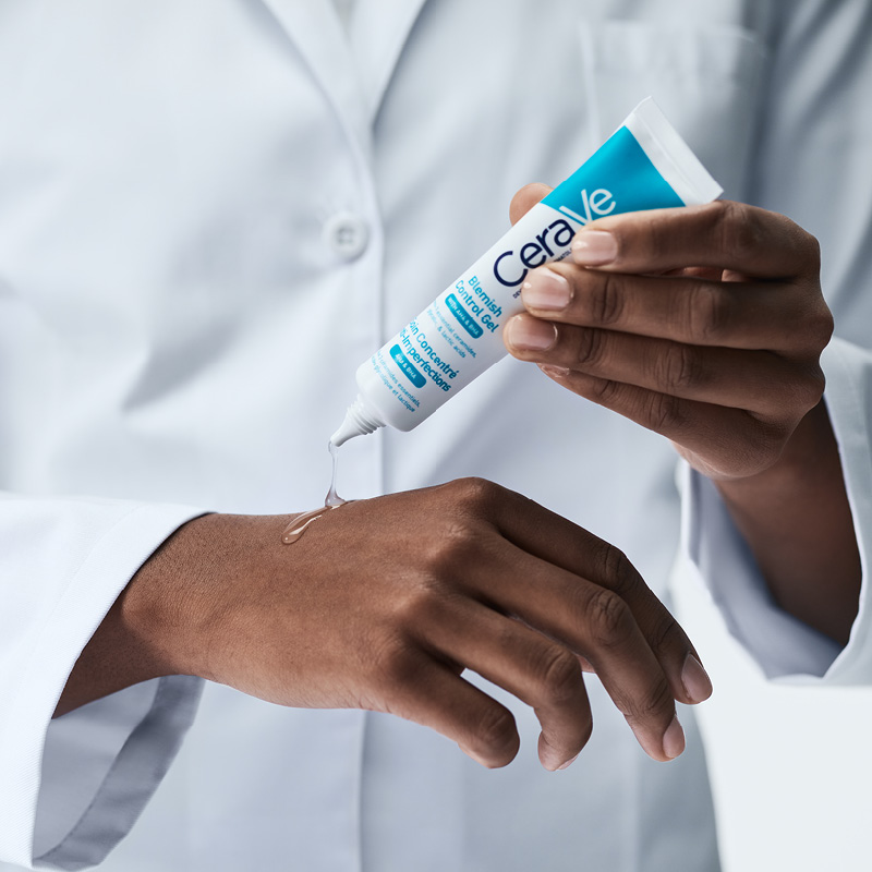 En person som applicerar CeraVes Blemish Control Gel på sin hand. Personen bär en vit labbrock och håller en tub med gel, som är vit med blå och svart text. Bakgrunden är ljusgrå.