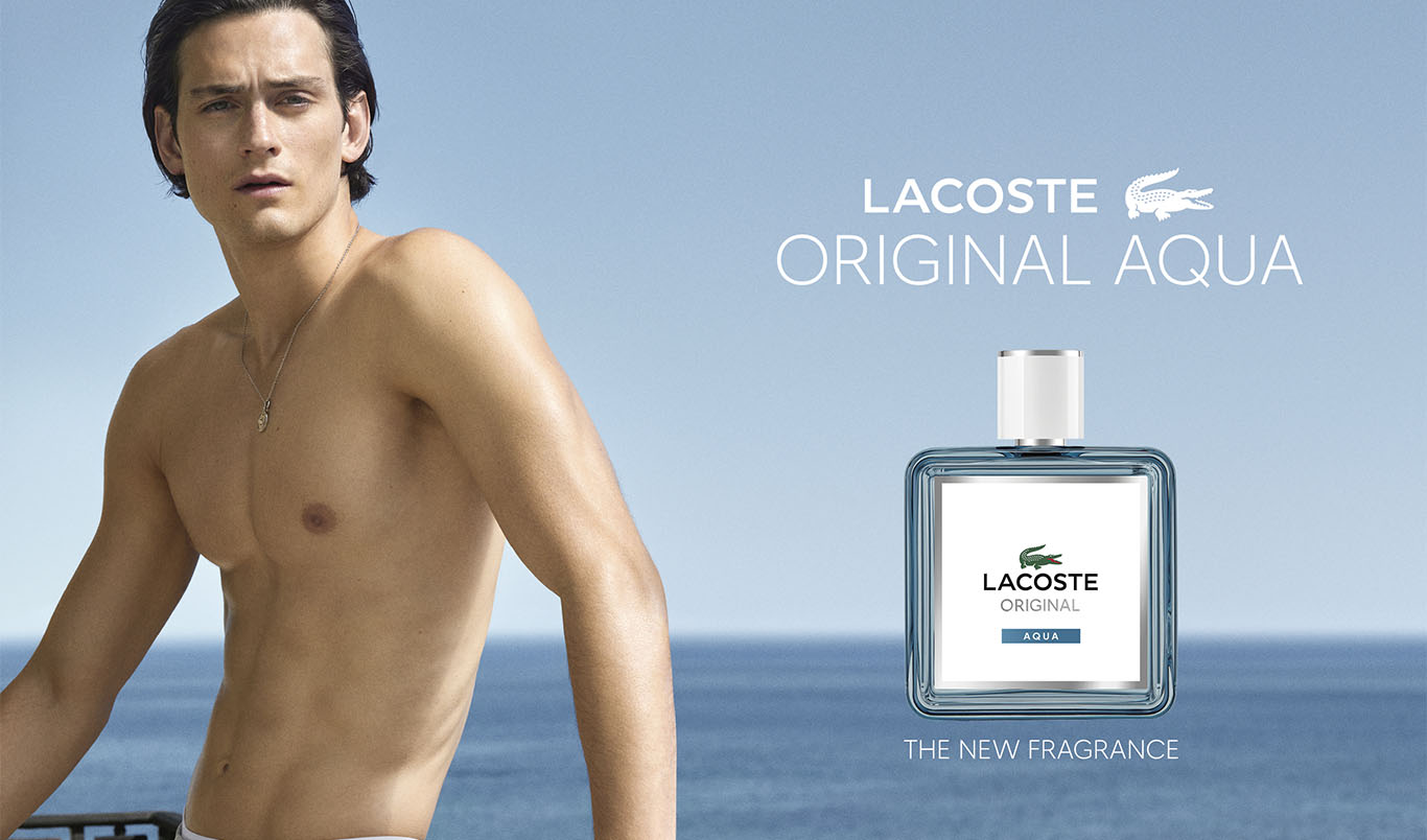 Paljas ylävartalo meren äärellä sekä vieressä hajuvesipullo, jossa teksti “LACOSTE ORIGINAL AQUA” ja “THE NEW FRAGRANCE”. Pullossa näkyy krokotiililogo ja teksti “LACOSTE ORIGINAL AQUA”.