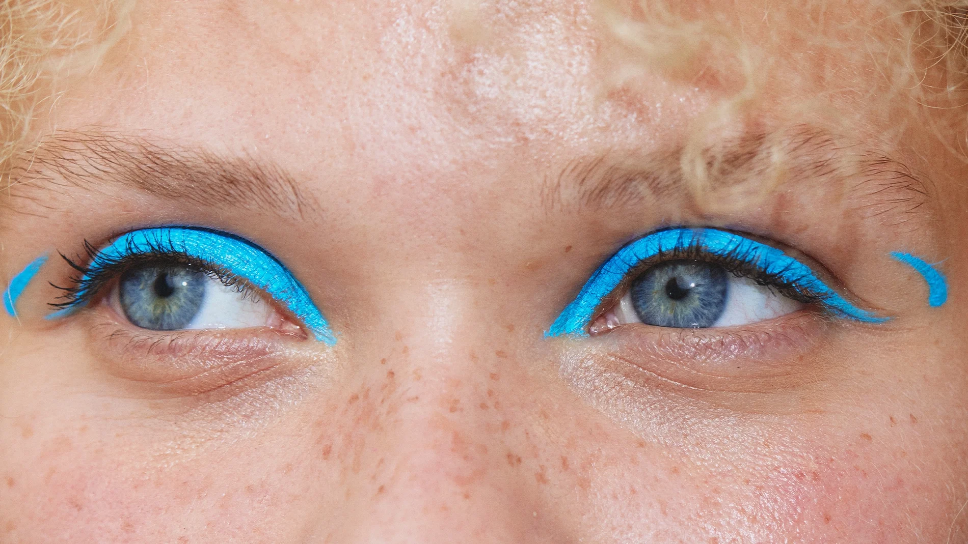 Närbild av ögon med intensivt turkosblå eyeliner längs övre ögonlocken, utdragen i grafiska vingar vid ögonvrårna. Små turkosblå detaljer syns utanför båda ögonen.