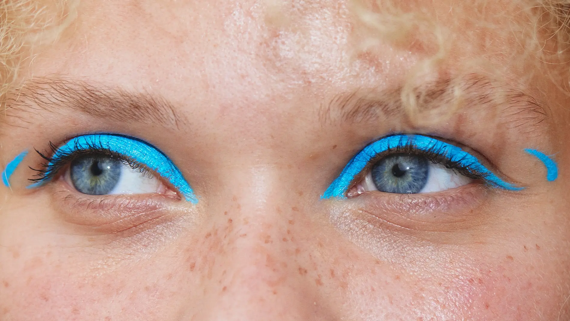Lähikuva silmistä, joiden yläluomilla on intensiivisen turkoosinsininen eyeliner, joka jatkuu graafisina siipinä silmäkulmissa. Molempien silmien ulkopuolella näkyy pieniä turkoosinsinisiä yksityiskohtia.