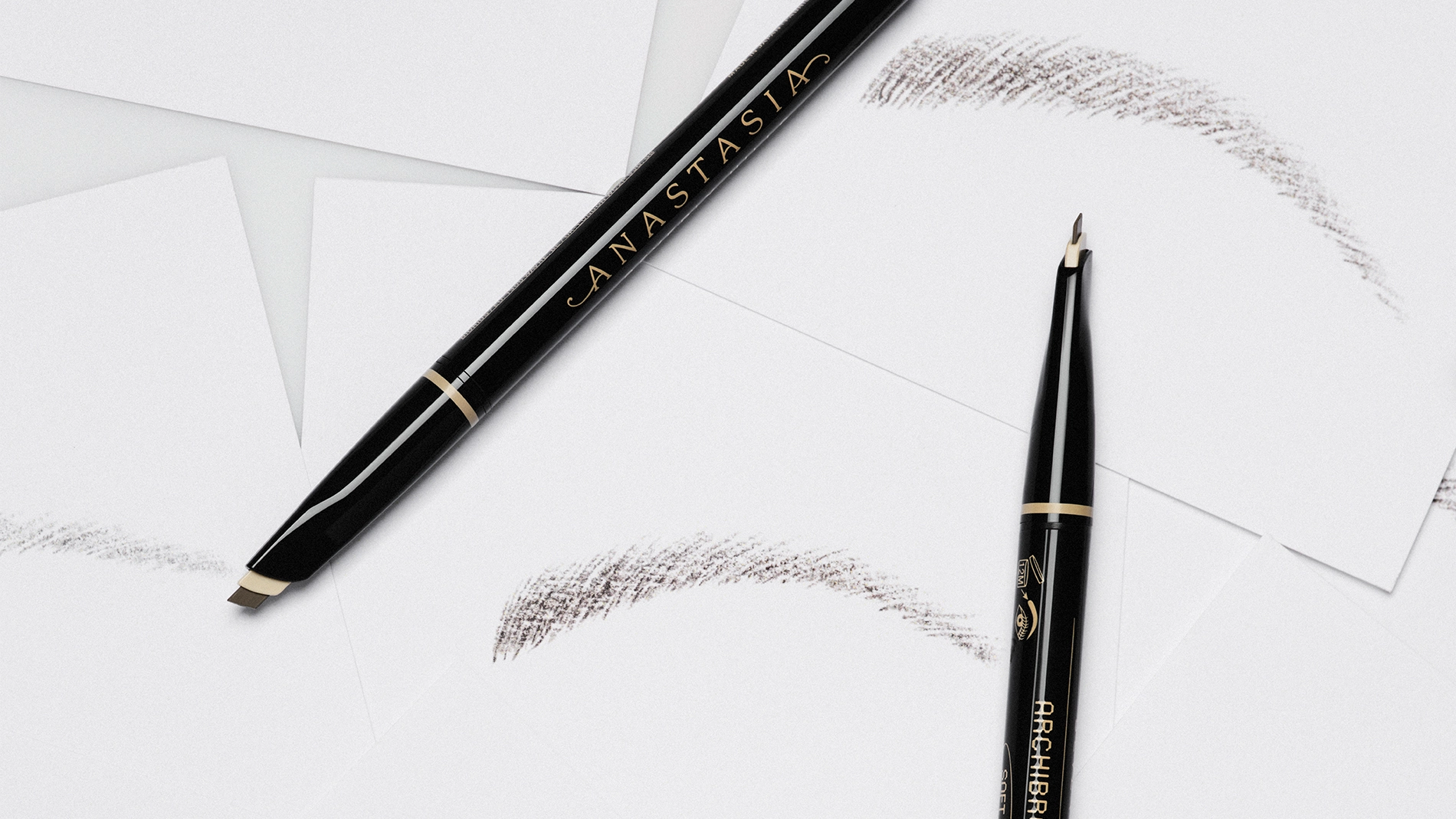 Svart øyenbrynsblyant fra Anastasia Beverly Hills med teksten "ANASTASIA" og "BROW WIZ", sammen med en spooliebørste og flere strøk av brynfarge på hvit bakgrunn.