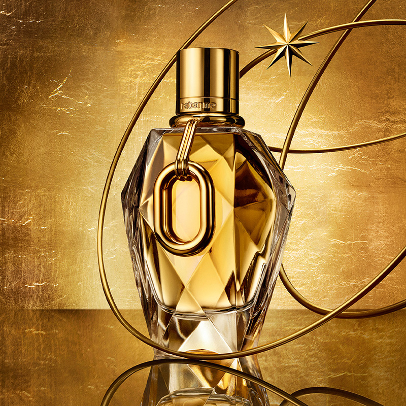 En gullfarget parfymeflaske med en geometrisk design står mot en bakgrunn med gulltoner. En stjerne og sirkulære linjer omgir flasken.