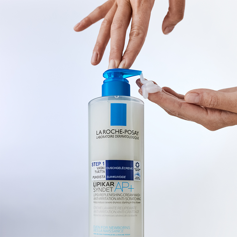 En hand pumpar ut en liten mängd av La Roche-Posay Lipikar Syndet AP+ duschgelkräm från en stor flaska med blå pump. Bakgrunden är ljus och minimalistisk, vilket framhäver produktens dermatologiska egenskaper.