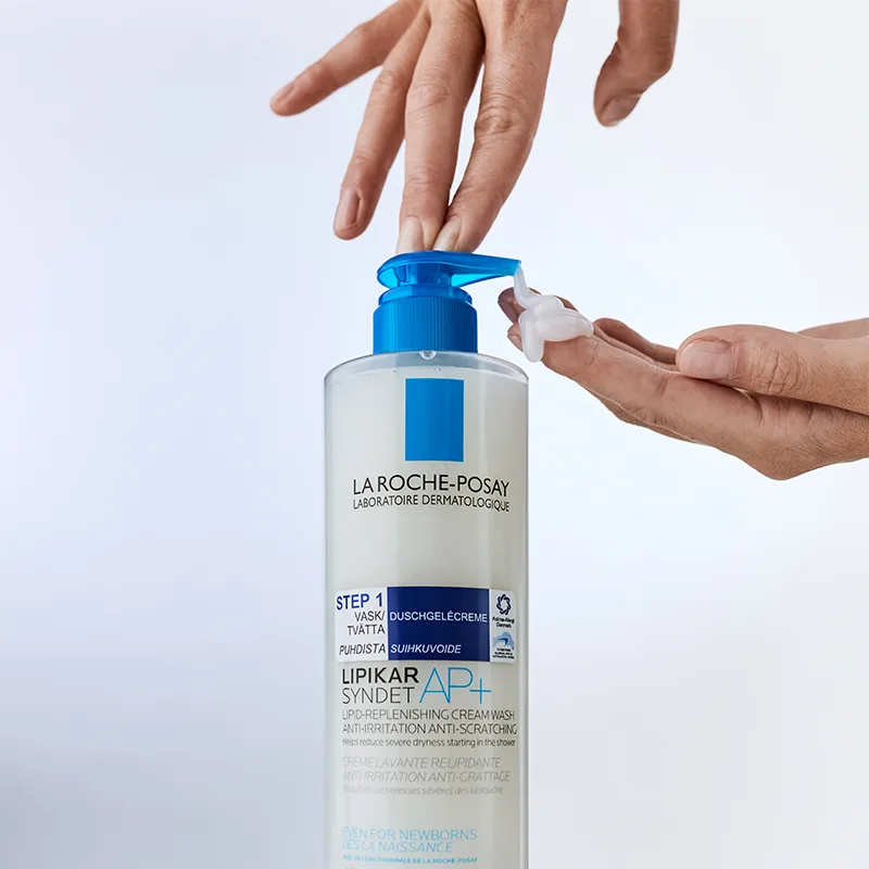 En hand pumpar ut en liten mängd av La Roche-Posay Lipikar Syndet AP+ duschgelkräm från en stor flaska med blå pump. Bakgrunden är ljus och minimalistisk, vilket framhäver produktens dermatologiska egenskaper.