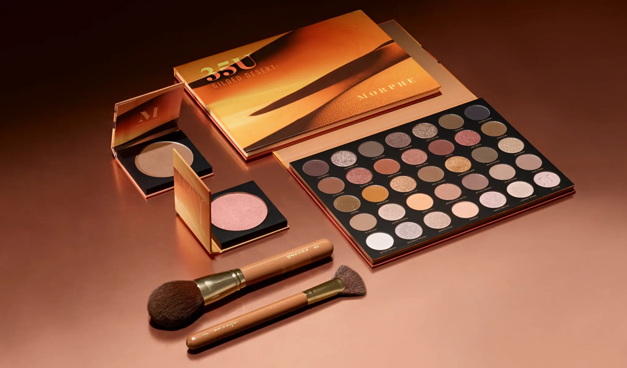 Morphe 35U Gilded Desert-øyenskyggepalett med et utvalg av varme nyanser, sammen med to sminkebørster og to highlightere i gull- og rosatoner, mot en kobberfarget bakgrunn.