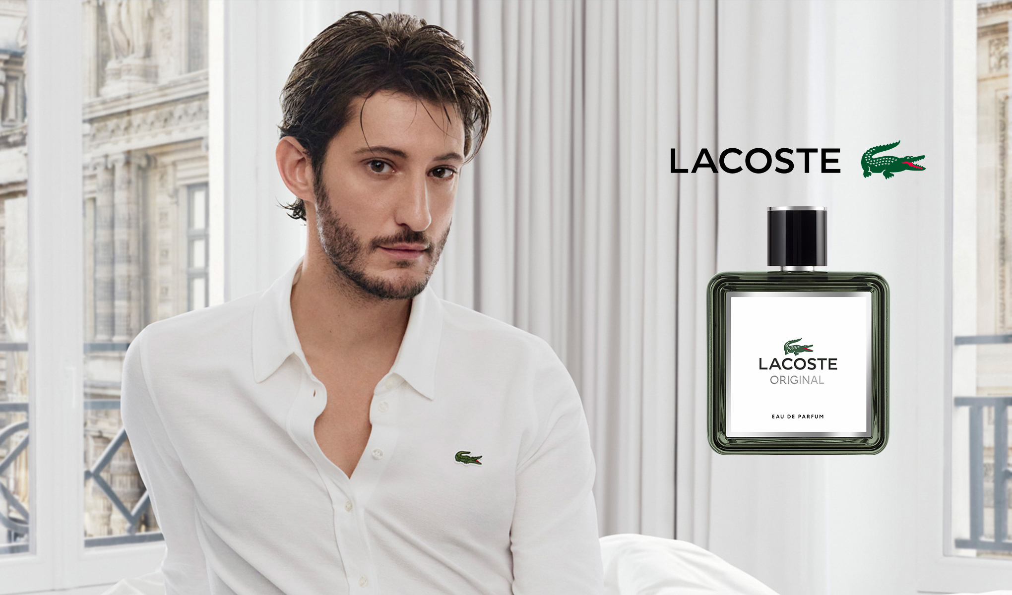 Valkoiseen Lacoste-paitaan pukeutunut henkilö istuu ikkunan edessä, josta on näkymä klassisen arkkitehtuurin rakennukseen. Henkilön vieressä on pullo Lacoste Original Eau de Parfumia. Hajuvesipullon yläpuolella on teksti ”Lacoste” ja Lacosten logo, jossa on vihreä krokotiili.