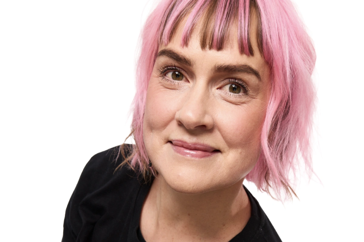 Porträtt av en person med kort rosa hår och naturlig makeup mot ljus bakgrund.
