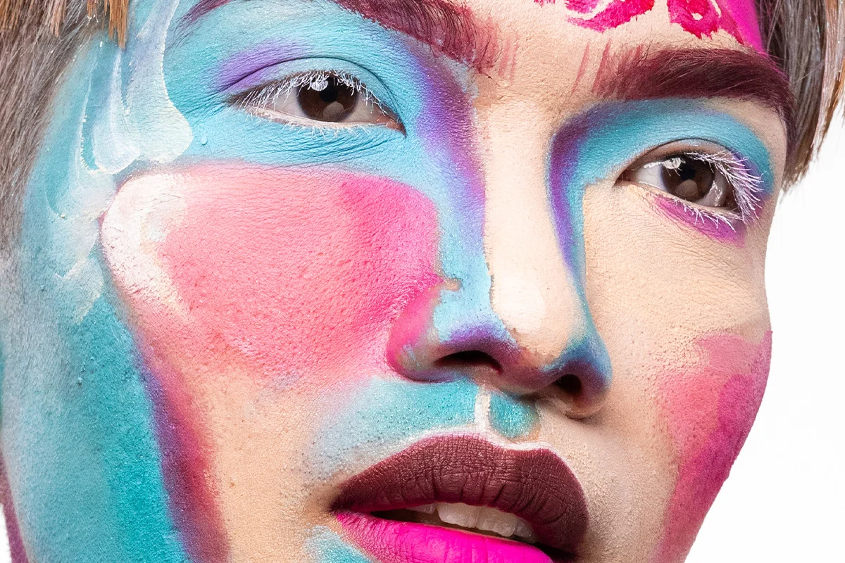 Extrem närbild av flerfärgad ansiktsmålning där blå, rosa och lila nyanser möts runt ögonen och kinderna, med vita fransar och tvåfärgade läppar som förstärker en ”living canvas”-inspirerad makeup.