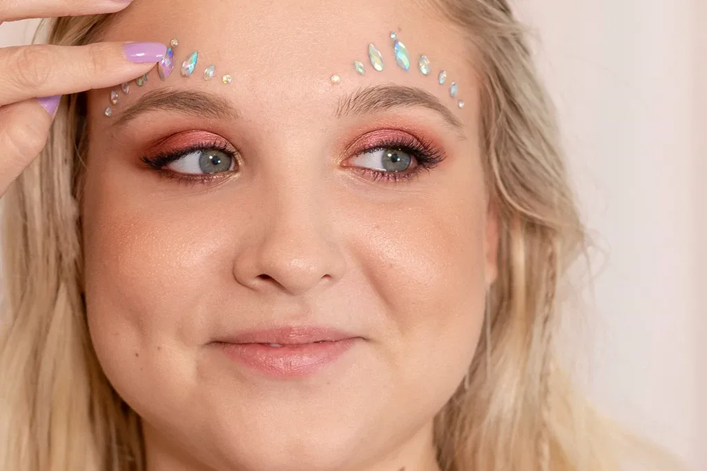 Blond kvinne med glitrende rosa øyenskygge og glødende sminke påfører rhinestones på toppen av øyenbrynene.