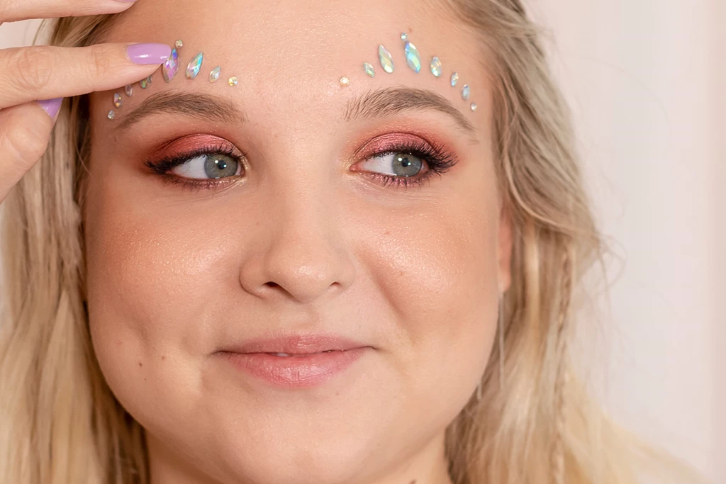 Vaalea nainen, jolla on hohtavan ruusuinen luomiväri ja hehkuva meikki, levittää strasseja kulmakarvojensa päälle.