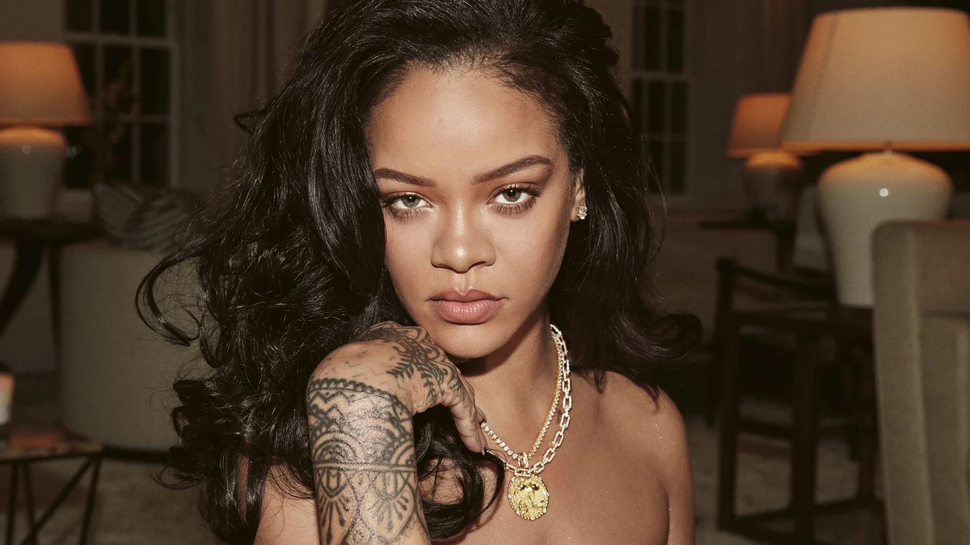 Rihanna tittar in i kameran iklädd halsband och visar hand med tatueringar. Sitter i vardagsrumsmiljö.