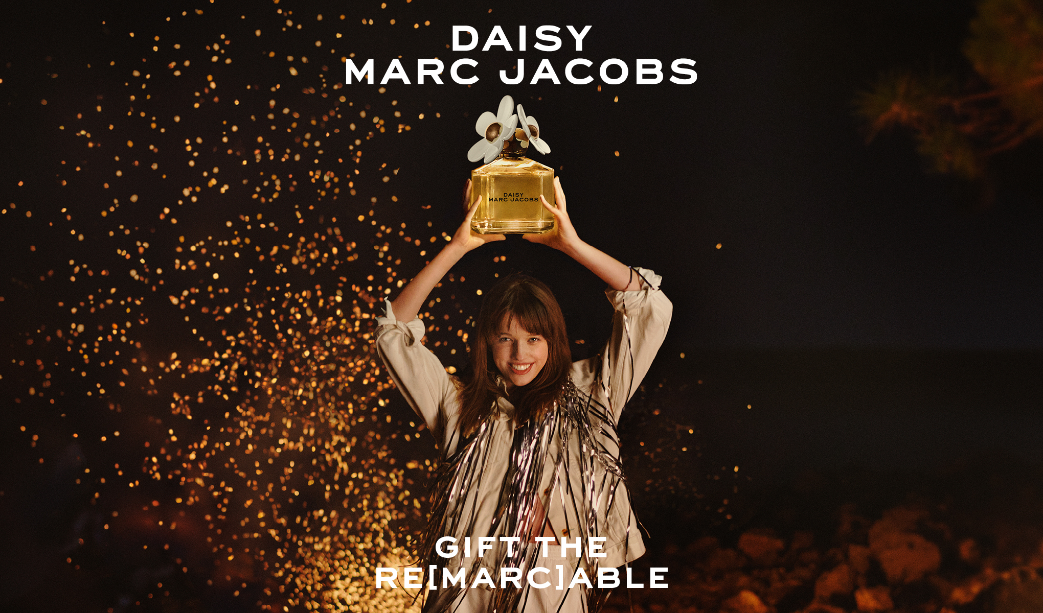 Henkilö pitää ylhäällä suurta Daisy Marc Jacobs -hajuvesipulloa tähtitaivaan alla, ympärillään kultaisia kipinöitä. Teksti "DAISY MARC JACOBS" yläosassa ja "GIFT THE RE[MARC]ABLE" alaosassa.