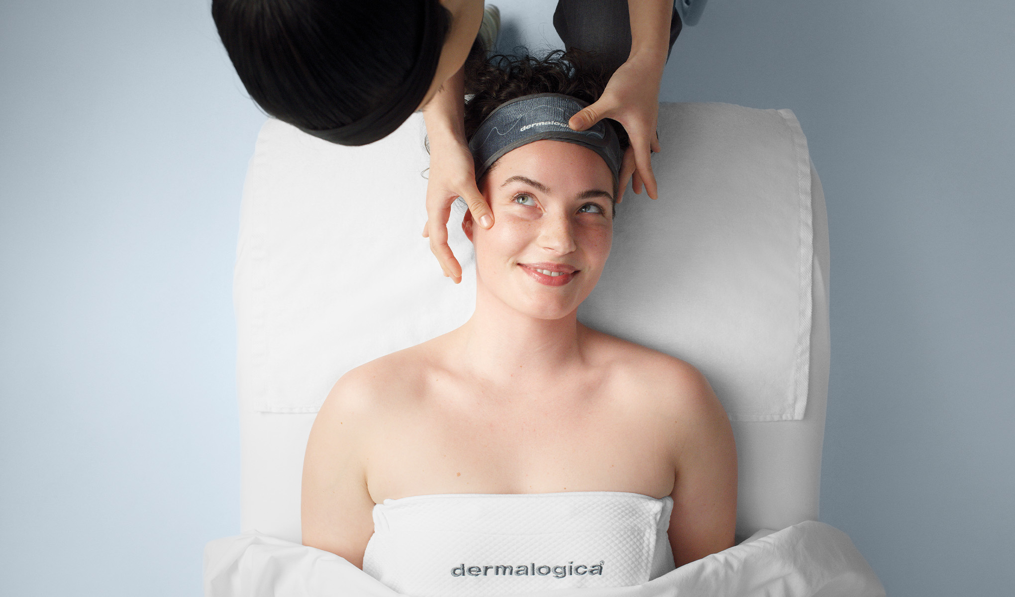 En person ligger på en behandlingsbänk med en vit handduk över sig, märkt med "Dermalogica". En terapeut med mörkt hår och grå kläder utför en ansiktsbehandling på personen. Bakgrunden är ljusblå och ger en lugn och avslappnad atmosfär.