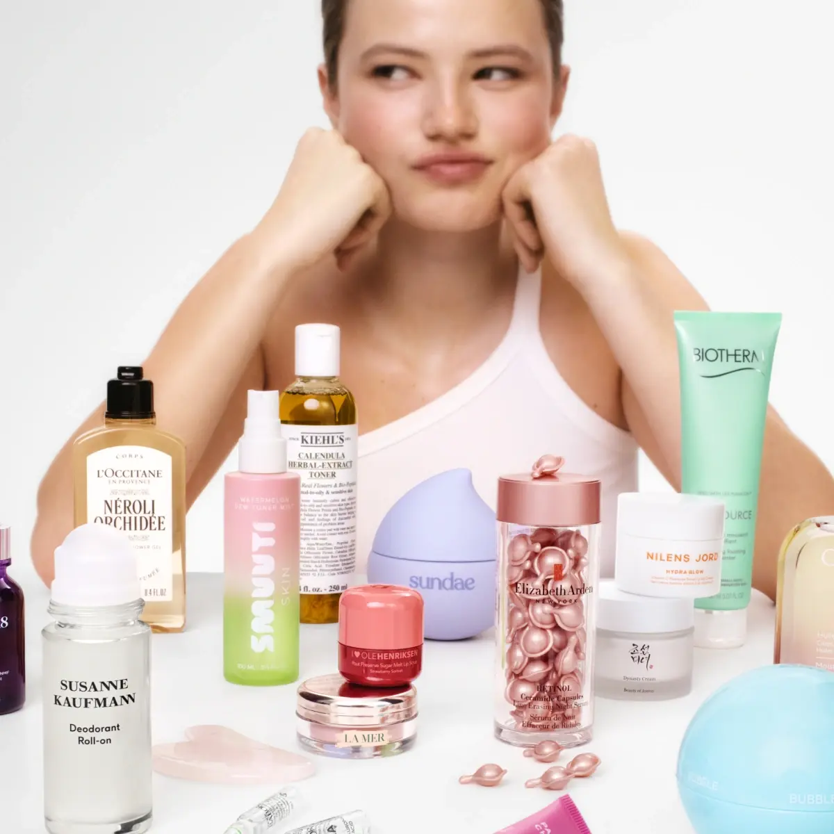 Person sitter bakom ett urval av hudvårdsprodukter från olika varumärken, inklusive Kiehl’s, Biotherm, La Mer och Elizabeth Arden, placerade på bord.