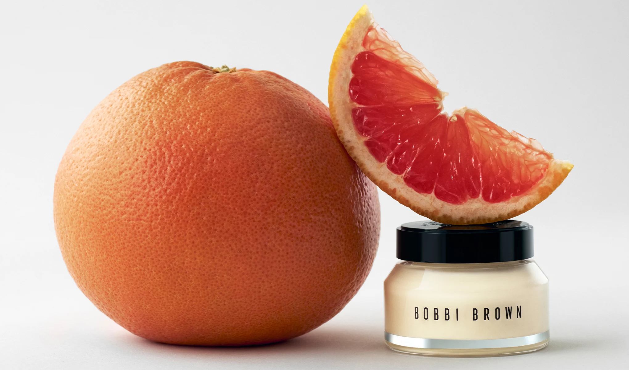 Bobbi Brown ansiktskrem i en glasskrukke med svart lokk står ved siden av en hel grapefrukt og en grapefruktskive mot en lys bakgrunn. Synlig tekst: "BOBBI BROWN" på krukken.