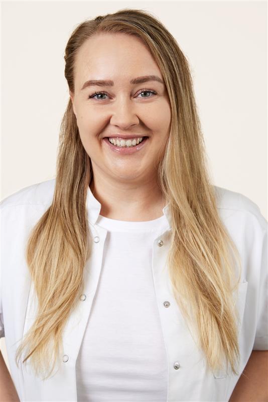 Porträtt av en leende person med långt blont hår och vit arbetsrock över vit topp mot ljus bakgrund. Personen bär naturlig makeup med lätt glow och definierade bryn.