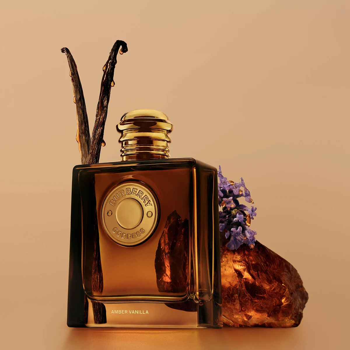 Burberry Goddess Amber Vanilla -hajuvesi tumman meripihkanvärisessä pullossa, jossa on kullanvärisiä yksityiskohtia, vaniljatankojen, violettien kukkien ja meripihkanvärisen kiven ympäröimänä. Pullossa näkyvät tekstit ”BURBERRY GODDESS” ja ”AMBER VANILLA”.