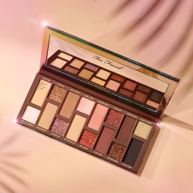 En øyenskyggepalett fra Too Faced med ulike farger og nyanser, åpnet og plassert på en rosa flate. Paletten har et speil.