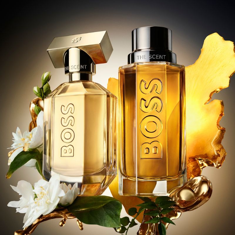 Kaksi Hugo Bossin 'The Scent' hajuvesipulloa, taustalla kukkia ja kultaisia koristeita.