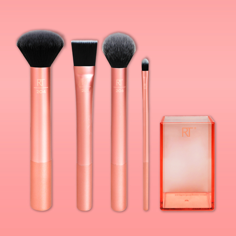 Fyra sminkborstar och en förvaringslåda från Real Techniques är arrangerade mot en rosa bakgrund. Borstarna har roséguldfärgade skaft och svarta borst. Förvaringslådan är transparent med en orange ton och har Real Techniques-logotypen på framsidan.