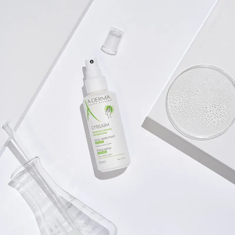 Vit sprayflaska från märket A-Derma med texten "Cytelium Drying Spray". Flaskan ligger på en vit yta tillsammans med ett genomskinligt lock, en laboratorieflaska och en petriskål med vätska. Produkterna är arrangerade på ett sätt som framhäver den kliniska och rena känslan. Bakgrunden är ljus och minimalistisk.