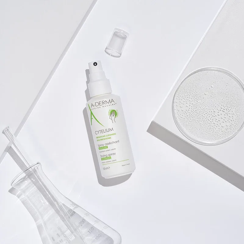 Vit sprayflaska från märket A-Derma med texten "Cytelium Drying Spray". Flaskan ligger på en vit yta tillsammans med ett genomskinligt lock, en laboratorieflaska och en petriskål med vätska. Produkterna är arrangerade på ett sätt som framhäver den kliniska och rena känslan. Bakgrunden är ljus och minimalistisk.