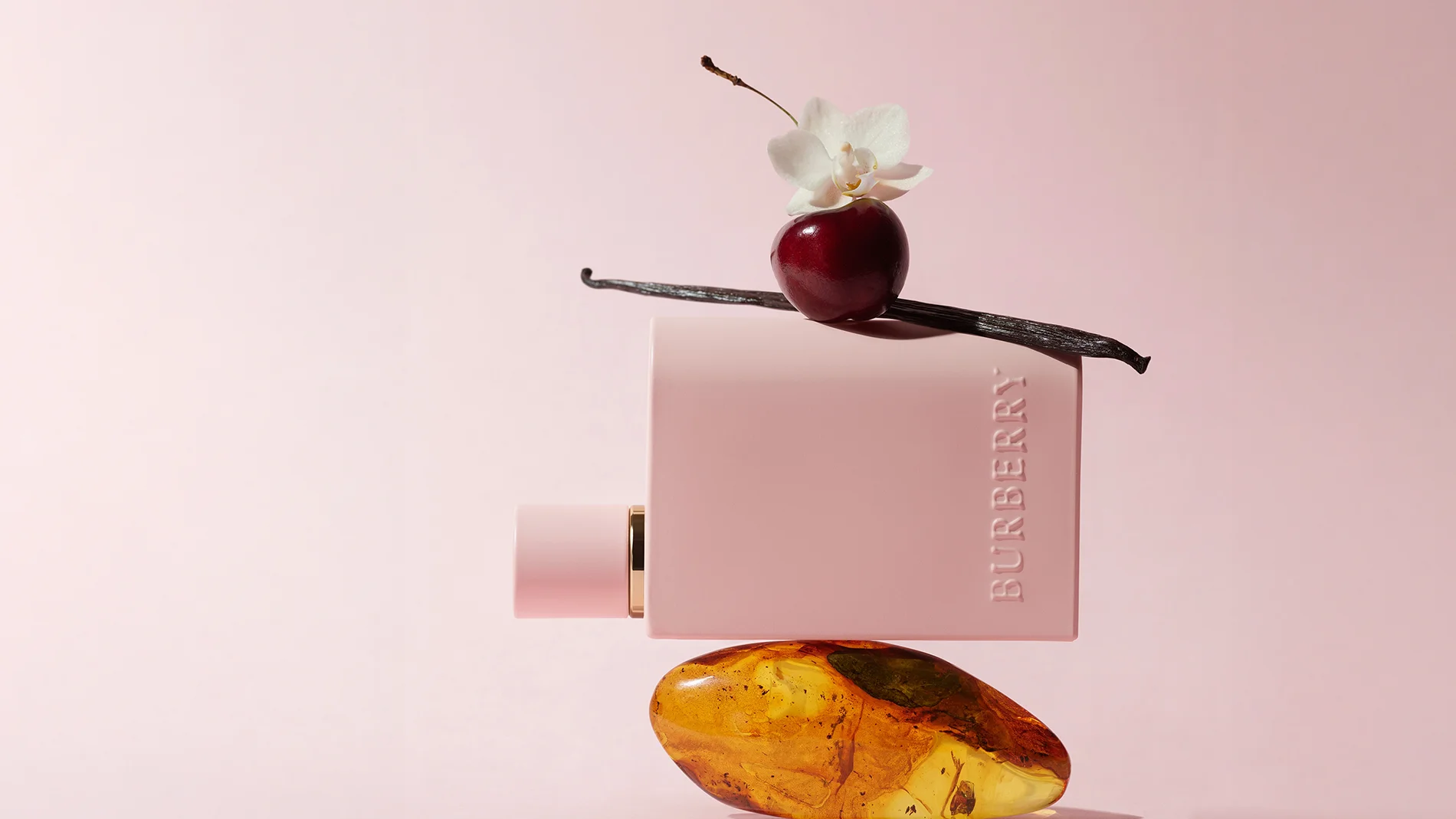 En rektangulær, lyserosa flaske med teksten «BURBERRY» liggende horisontalt. Flasken balanserer på en ravfarget stein. Oppå flasken ligger en mørk vaniljestang, et rødt bær og en hvit blomst. Bakgrunnen er ensfarget i en myk rosa tone.