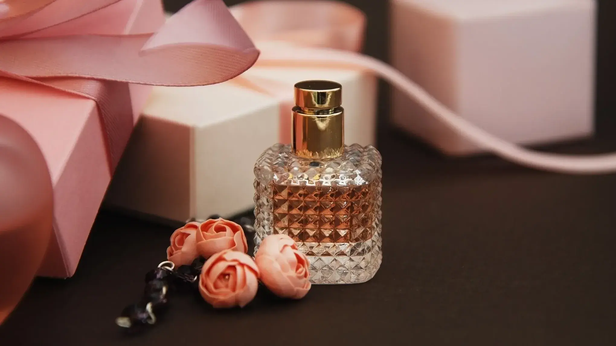 Parfymeflaske med gullfarget kork omgitt av små roser og gaveesker med rosa bånd.