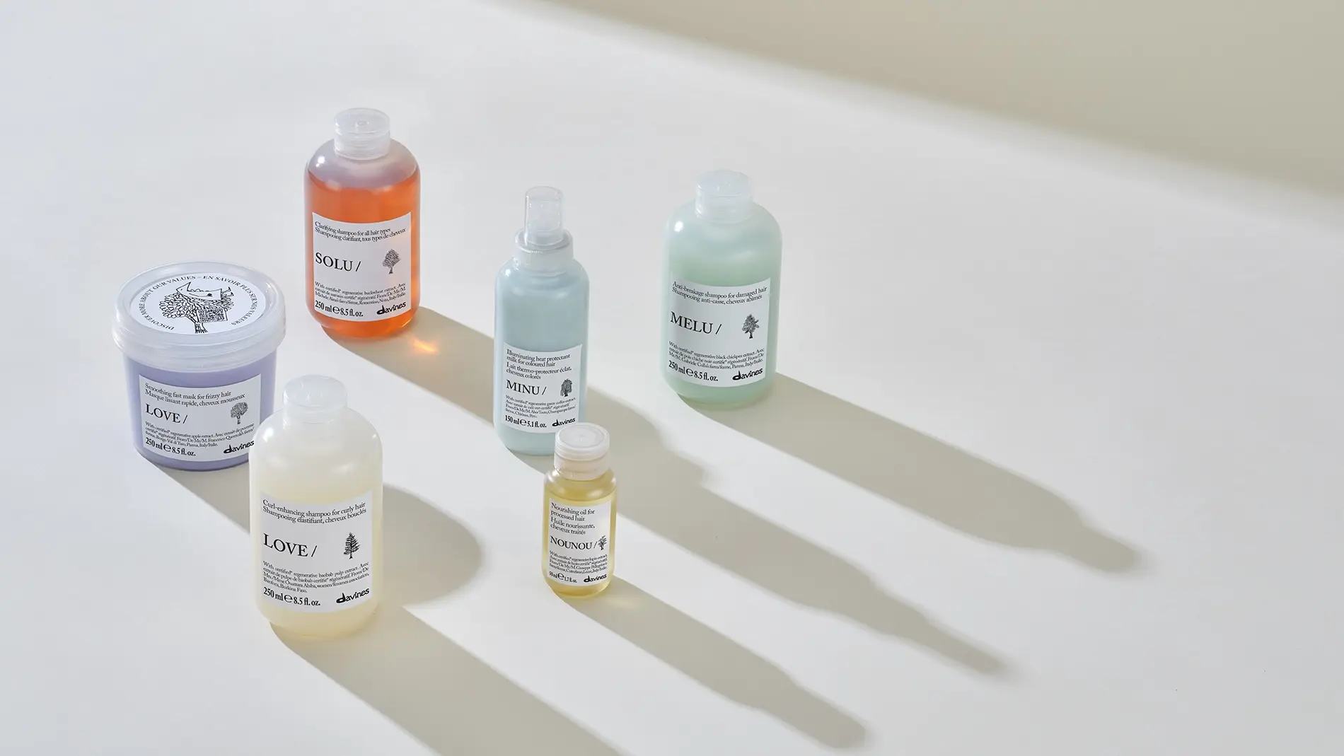 Davines hårvårdsprodukter från serierna LOVE, SOLU, MINU, MELU och NOUNOU står uppställda på vit yta med långa skuggor i mjukt ljus. Bilden visar schampo, hårmask, hårolja och värmeskyddsspray med produktnamn och Davines-logotyp synliga på förpackningarna.