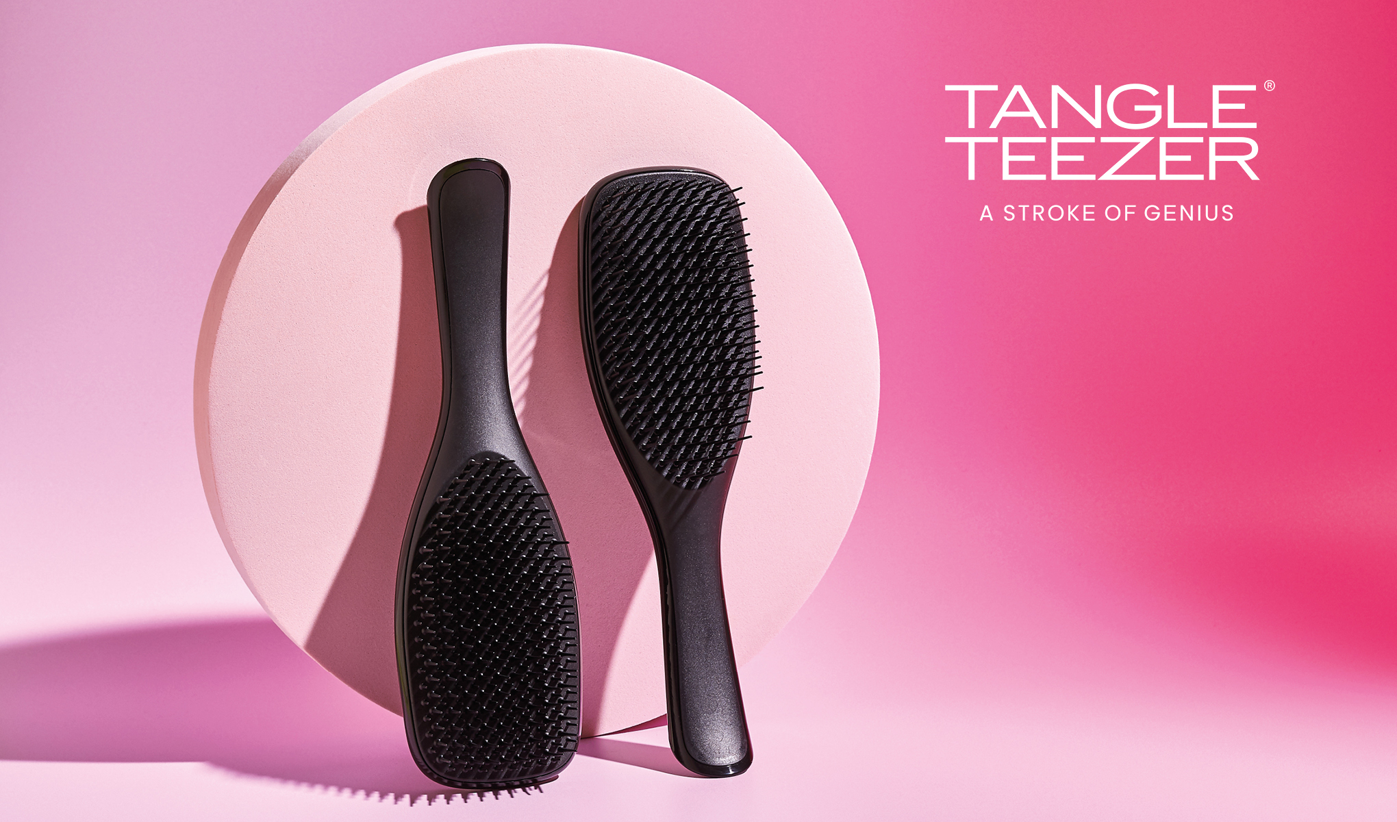 Kaksi mustaa hiusharjaa vaaleanpunaisella pohjalla, jossa on vaaleanpunainen pyöreä levy. Teksti ”Tangle Teezer A Stroke Of Genius” on sijoitettu oikeaan yläkulmaan. Harjissa on tyylikäs muotoilu.