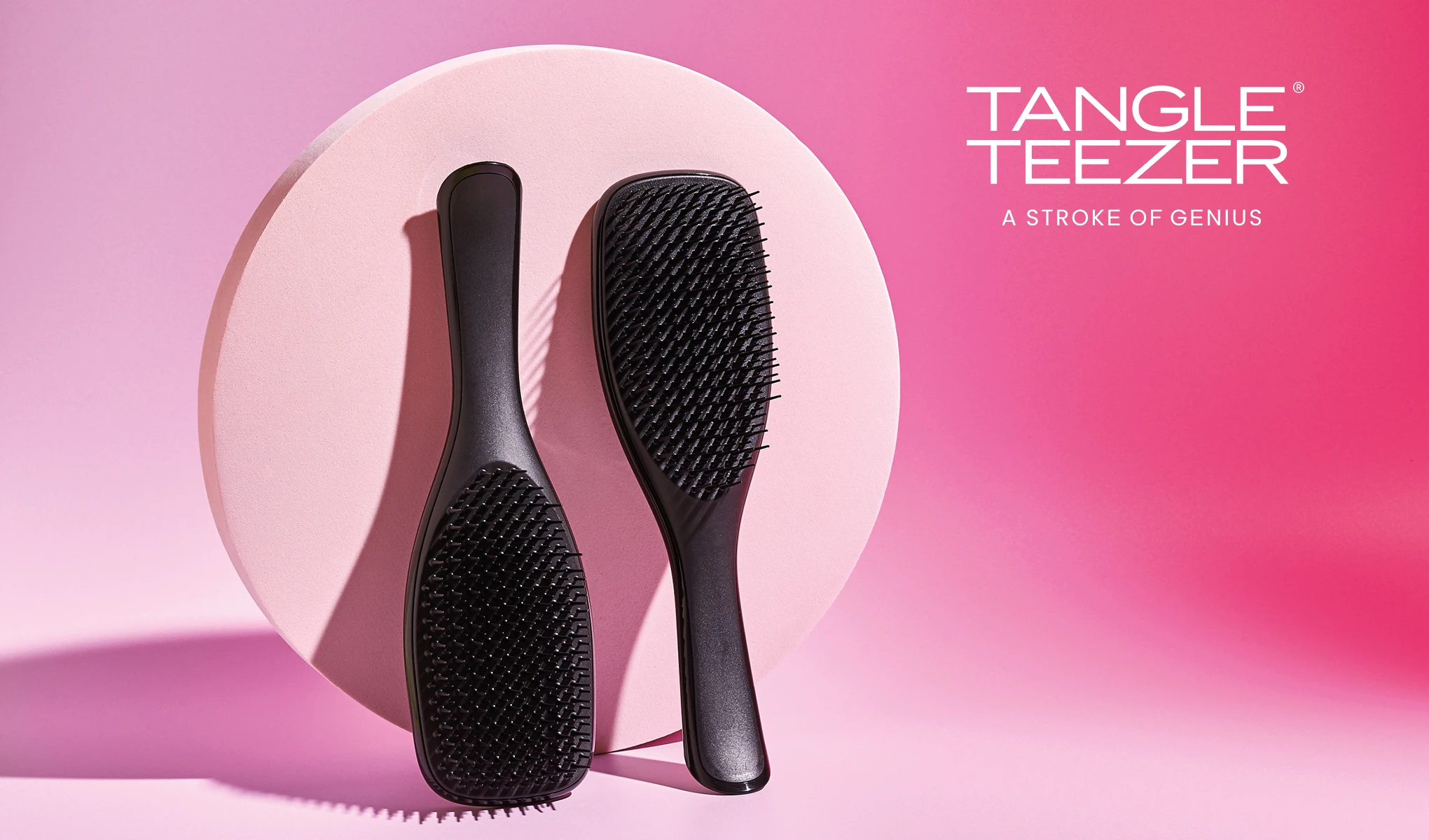 Kaksi mustaa hiusharjaa vaaleanpunaisella pohjalla, jossa on vaaleanpunainen pyöreä levy. Teksti ”Tangle Teezer A Stroke Of Genius” on sijoitettu oikeaan yläkulmaan. Harjissa on tyylikäs muotoilu.