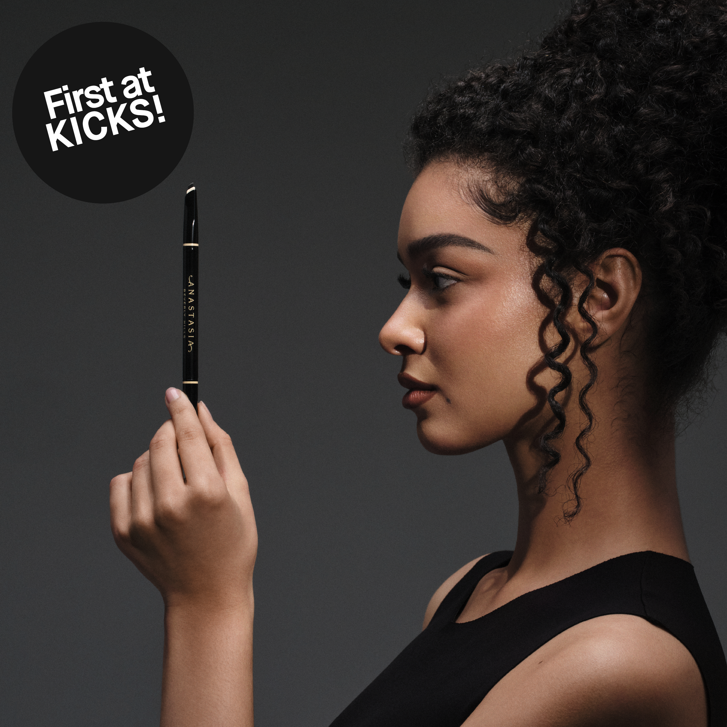 Nainen sivuprofiilissa pitelee mustaa kulmakynää kasvojensa edessä, tumma tausta ja teksti “First at KICKS!”.