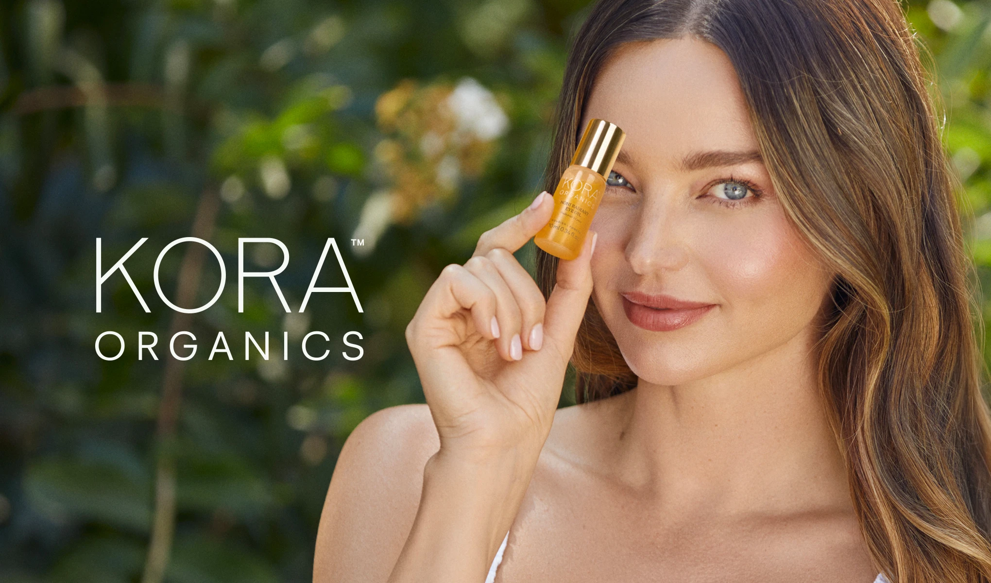 Henkilö pitää kädessään pientä oranssia pulloa vehreällä taustalla. Vasemmalla puolella on teksti "KORA ORGANICS".