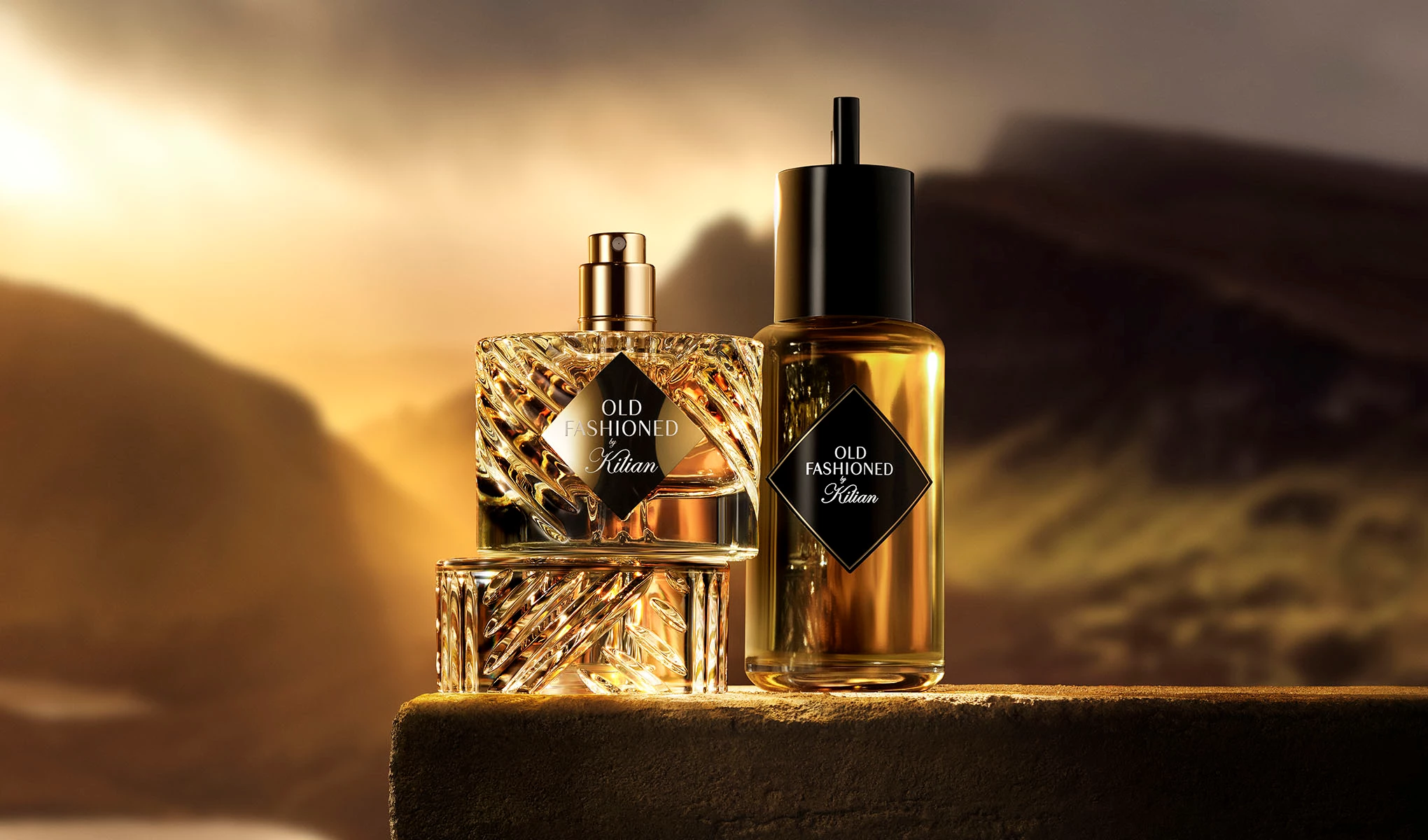 Två parfymflaskor med etiketten "Old Fashioned Kilian" står på en yta med en vacker, solnedgångsbelyst landskap i bakgrunden. Den ena flaskan har en elegant, kristallmönstrad design medan den andra är en enkel, cylindrisk flaska med svart kork.