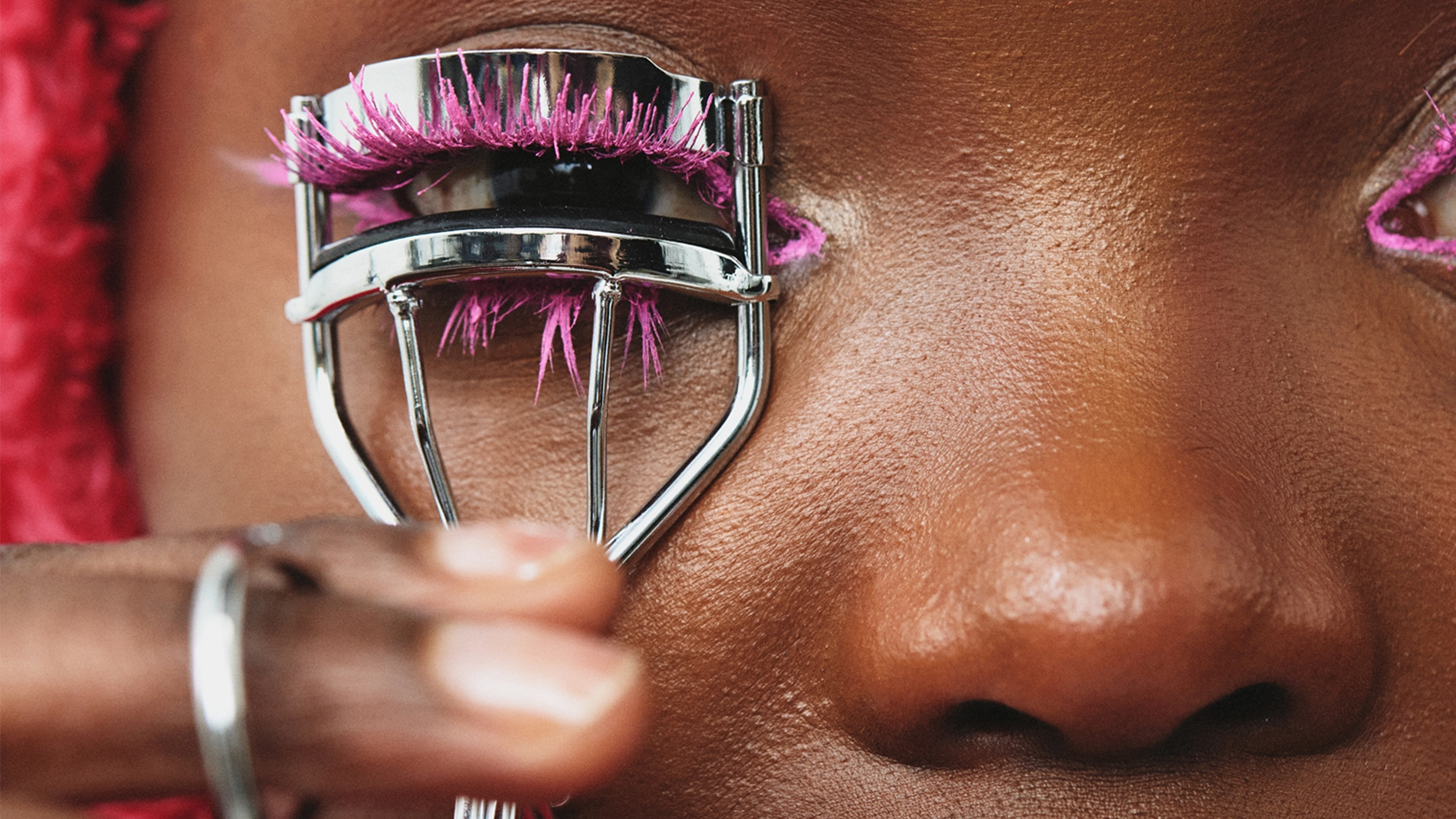 Nærbilde av en person som bruker en øyenvippebøyer med rosa kunstige øyenvipper. Personen har fyldige lepper med mørk lipgloss og bærer en rosa hodeplagg. Bildet fokuserer på øyenvippebøyeren og øyenvippene.