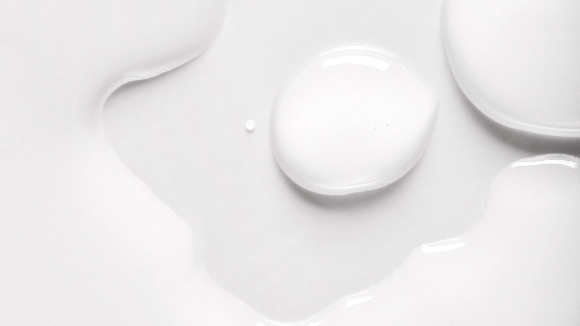 Närbild på vita vätskedroppar i olika former mot en ljus, slät yta med mjuka reflektioner.