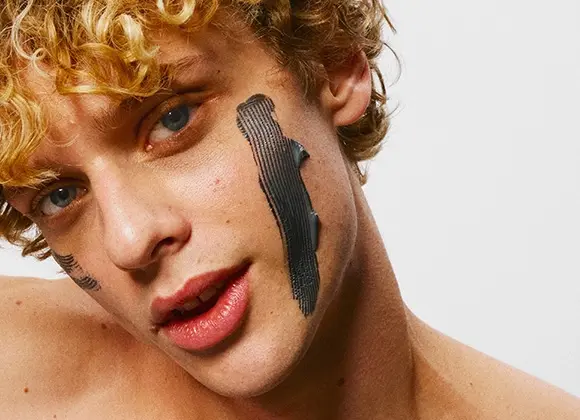 Närbild på person med lockigt blont hår och svart ansiktsmask applicerad i breda drag på kinderna.