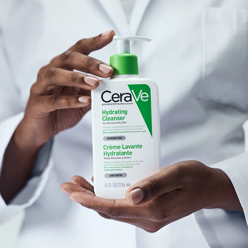 Två händer som håller en flaska av CeraVes Hydrating Cleanser. Flaskans etikett är vit med grön och svart text, och den har en grön pump. Bakgrunden är vit.