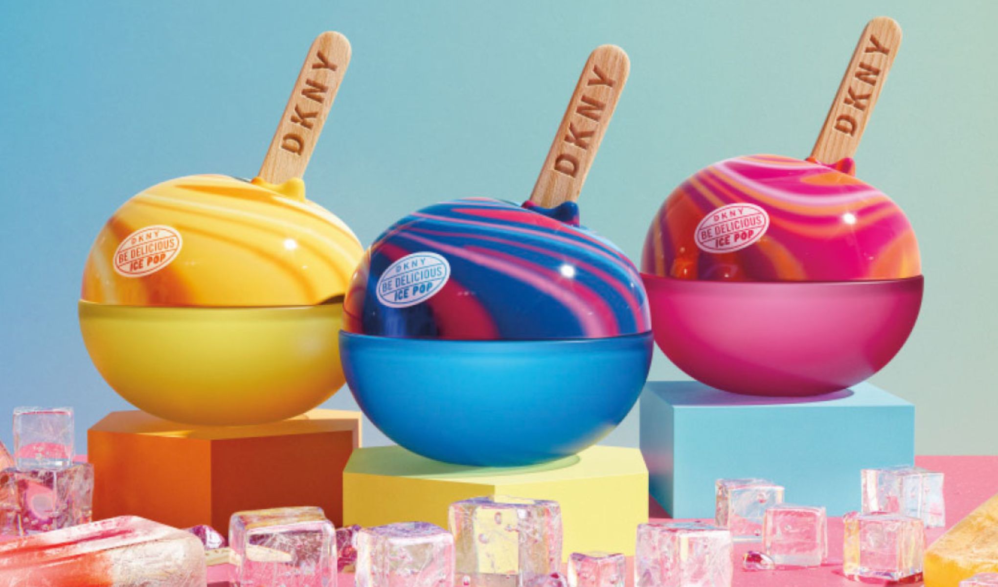 Tre färgstarka DKNY Be Delicious Ice Pop i glassformade förpackningar placerade på färgade podier, omgivna av isbitar mot en pastellfärgad bakgrund.