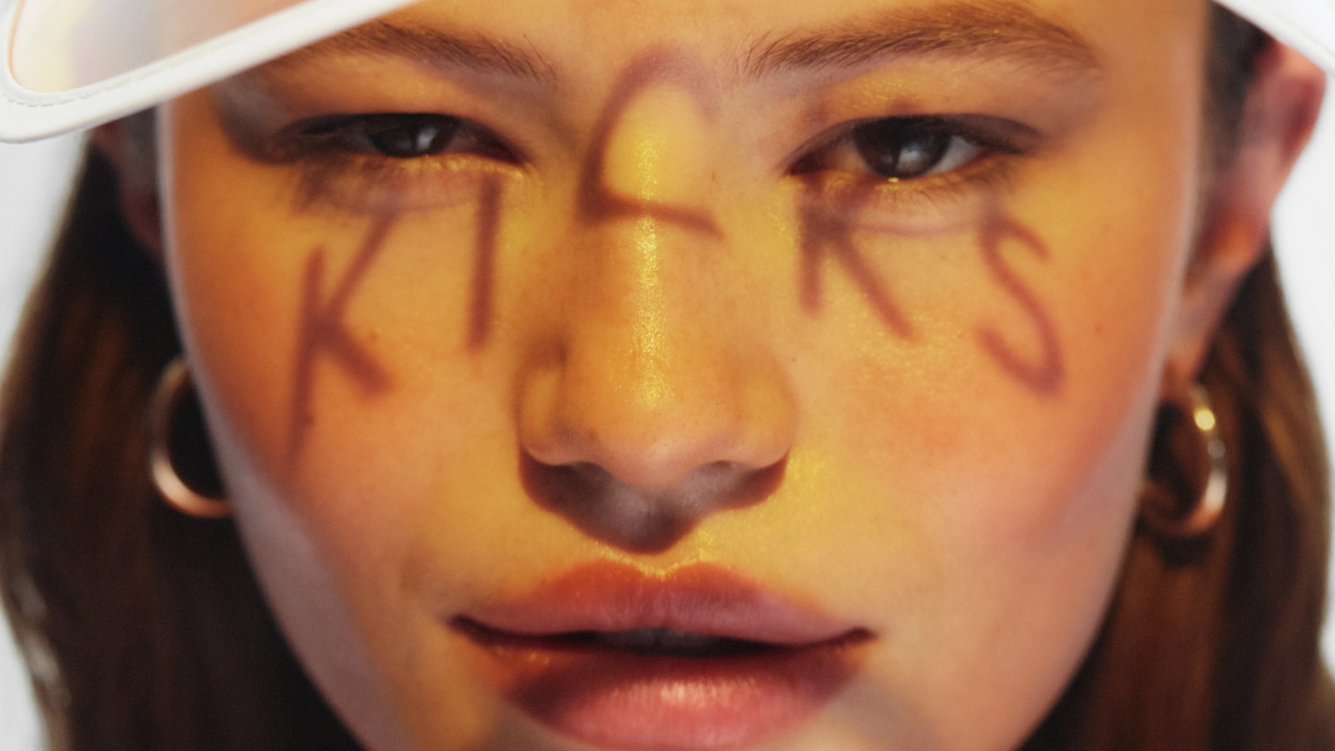 Nærbilde av ansikt med varm, gyllen sminke og glød i huden, med bokstavene "KICKS" malt over ansiktet. Fokus på øyne, hudfinish og kreativ makeup-look.