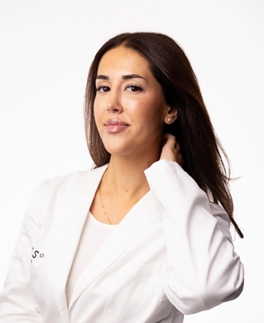 Porträtt av en person med naturlig makeup, glansigt läppstift och långt utsläppt hår mot ljus bakgrund. Personen bär en vit kavaj och smycken, vilket ger ett rent och professionellt uttryck kopplat till skönhet och hudvård.