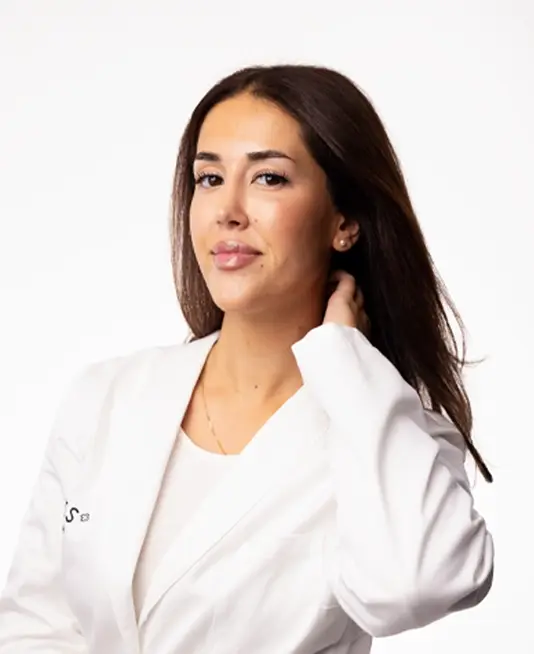 Porträtt av en person med naturlig makeup, glansigt läppstift och långt utsläppt hår mot ljus bakgrund. Personen bär en vit kavaj och smycken, vilket ger ett rent och professionellt uttryck kopplat till skönhet och hudvård.