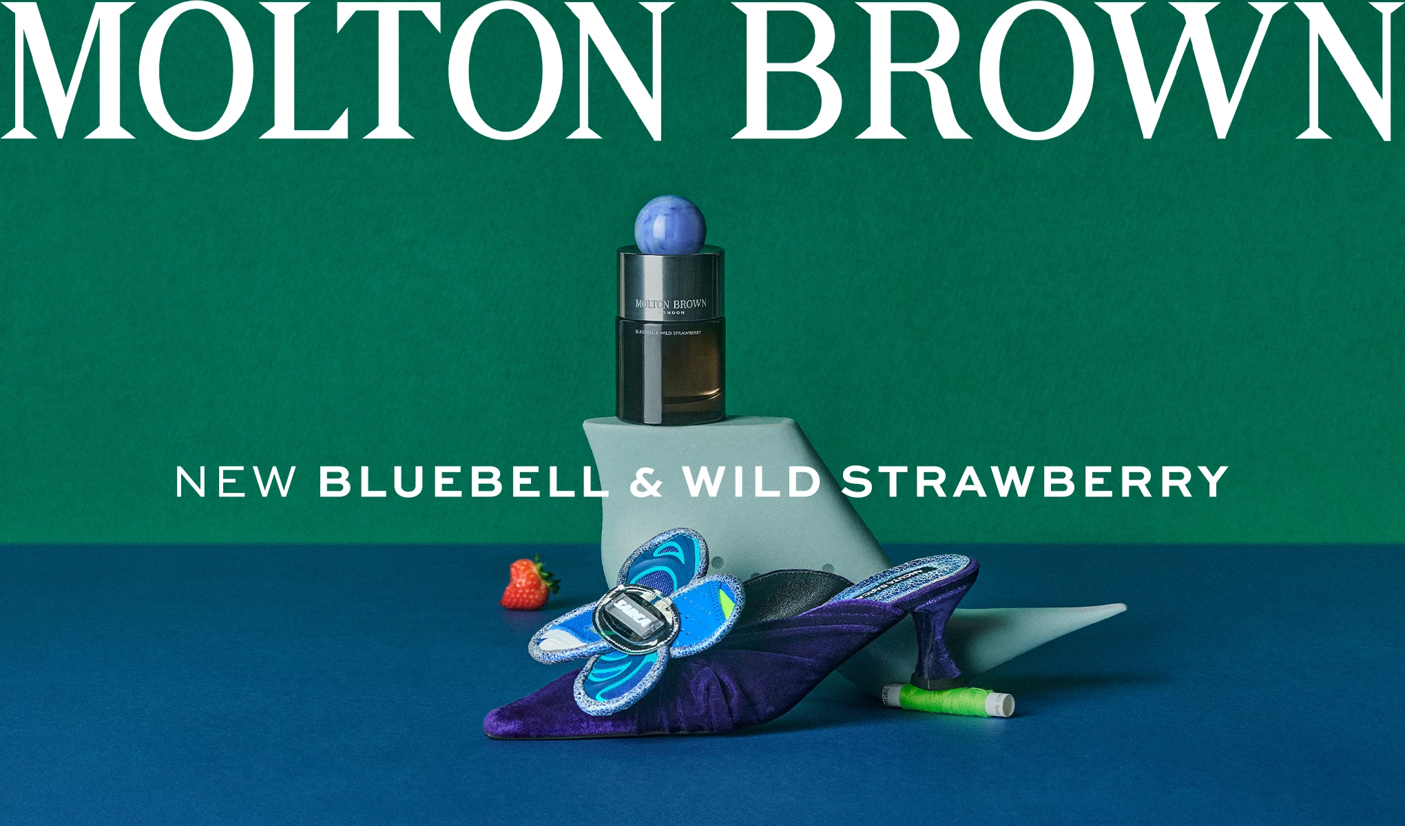 Molton Brownin hajuvesipullo, joka on sijoitettu vihreään geometriseen muotoon, sekä sininen ja violetti koristekenkä, jossa on perhonen. Taustalla on vihreä seinä ja sininen pinta sekä pieniä koriste-esineitä, kuten mansikka ja lankakela. Kuvan tekstissä lukee ”Molton Brown - New Bluebell & Wild Strawberry”.